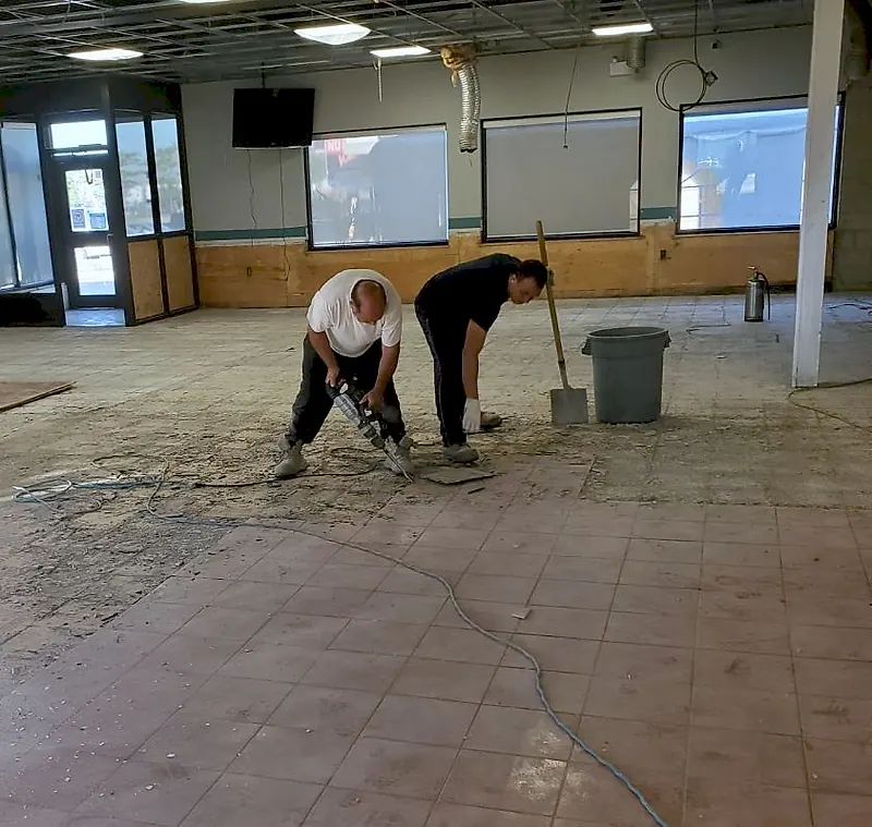 Two workers breaking out floor tile with a demolition hammer in an empty dining room
