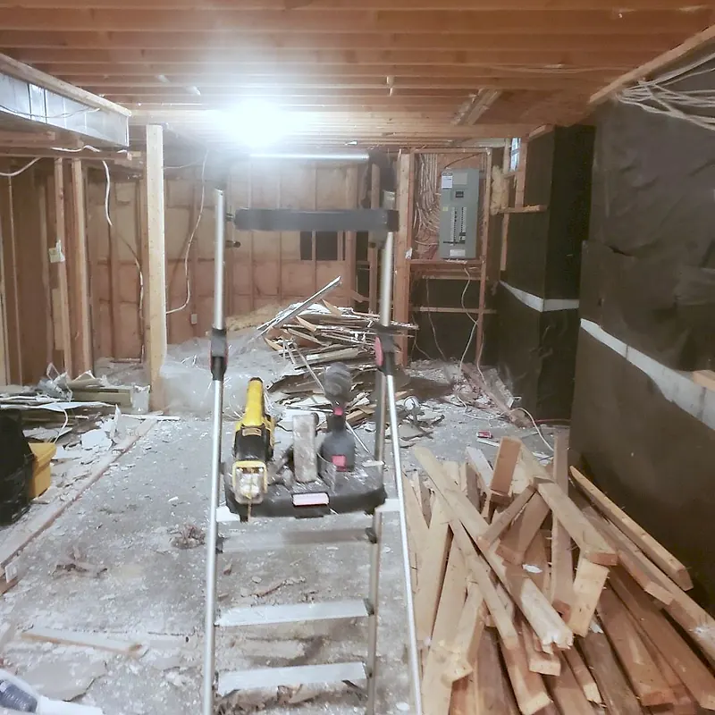 Basement stripped to the framing with lumber stacked and power tools on a stepladder