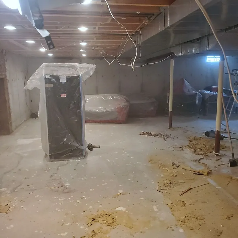 Empty basement with bare concrete walls and floor, a sofa and a tall cabinet wrapped in plastic sheeting