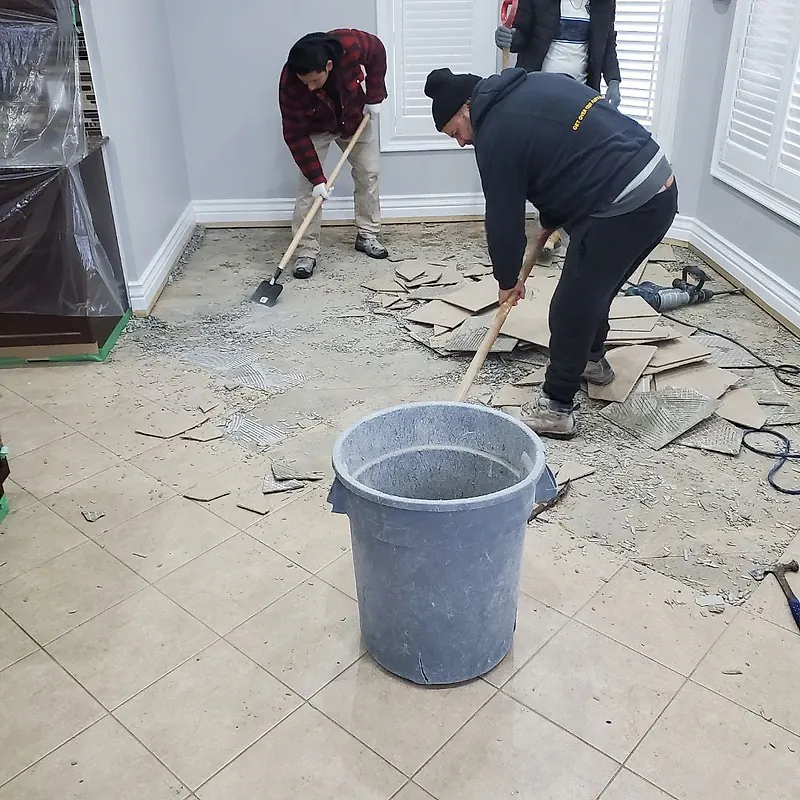 Workers lifting floor tile in a bright room, the furniture wrapped in plastic