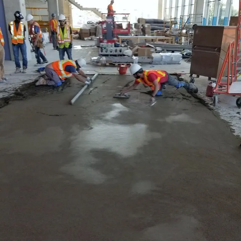 Two workers in hard hats and hi-vis vests kneeling on a wet mortar bed with a straightedge and a float in a large glazed atrium