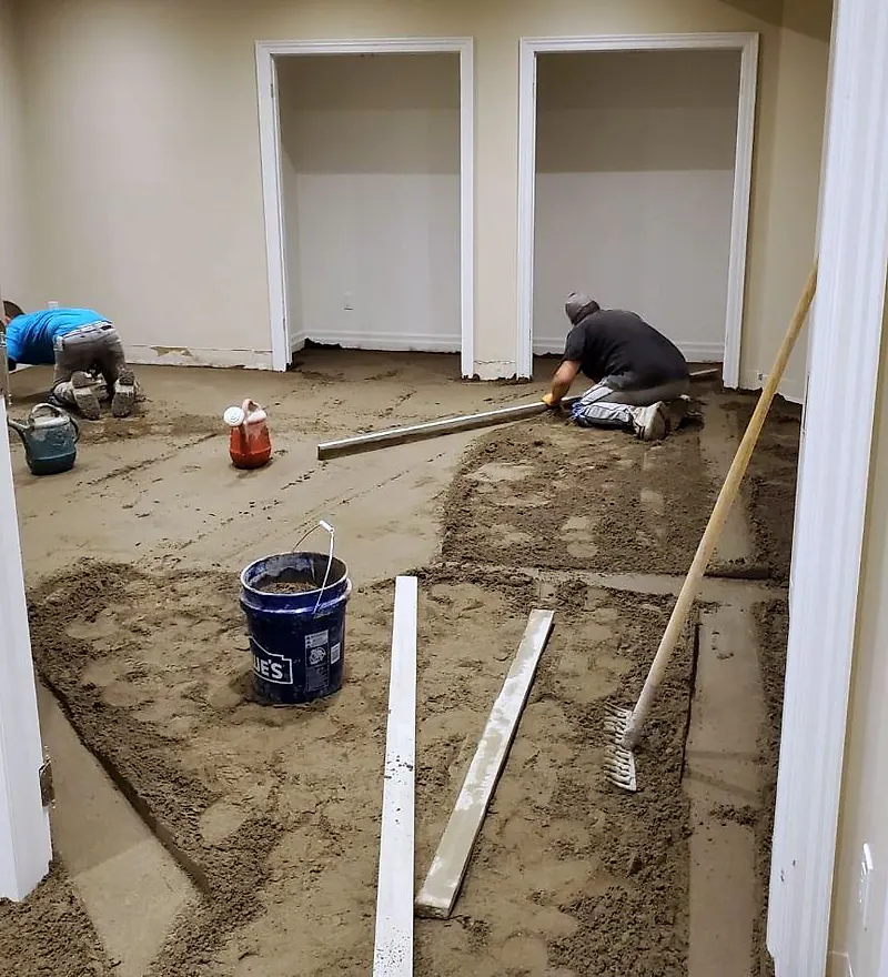 Two workers kneeling with straightedges spreading a mortar floor bed in a basement room with two closets
