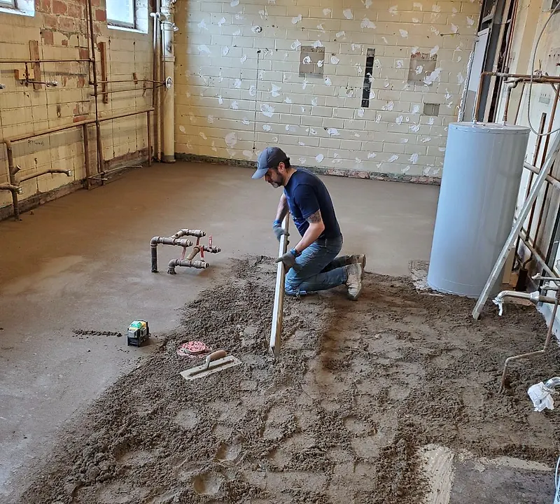 Worker kneeling with a straightedge spreading a mortar bed in an old block-walled basement with plumbing rough-ins