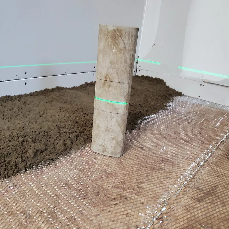 Wooden gauge stick with pencil marks and a green laser line across it, loose mortar and mesh on the floor