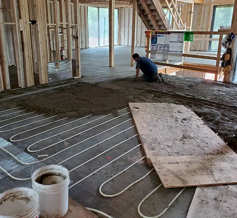 Worker kneeling and spreading mortar over white heating tubes in a wood-frame house under construction