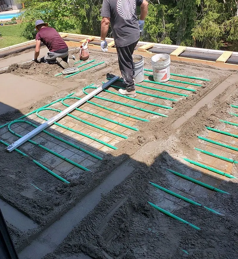 Green heating tubes on wire mesh on an outdoor terrace, with two workers placing mortar and a straightedge