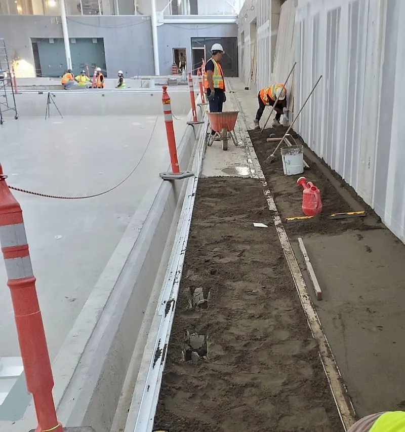 Narrow mortar bed being screeded between rails beside an indoor pool, with orange delineator posts and workers in hi-vis vests