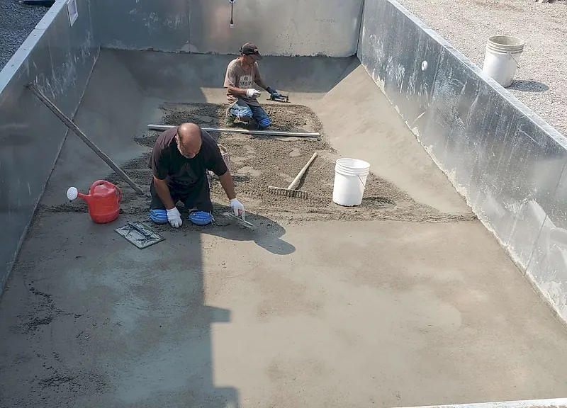 Two workers kneeling on a fresh mortar bed inside an empty pool, with a straightedge, buckets and a watering can