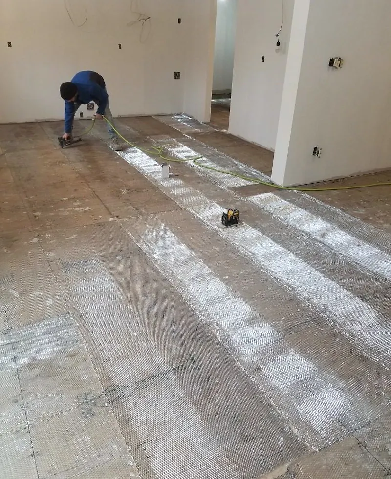 Worker fastening metal lath to a wood subfloor with an air stapler, a laser level on the floor