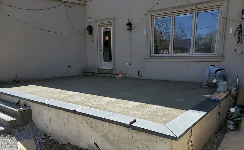 Raised concrete terrace with a fresh screeded mortar bed inside dark stone coping, a house wall behind