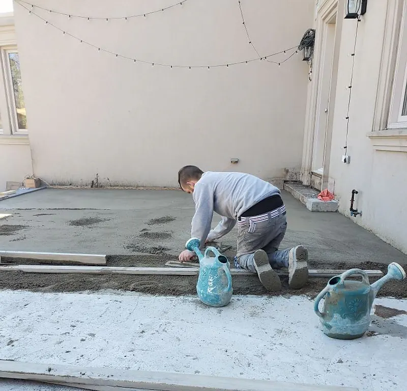 Worker kneeling between screed rails floating a mortar bed on a terrace, two watering cans beside him