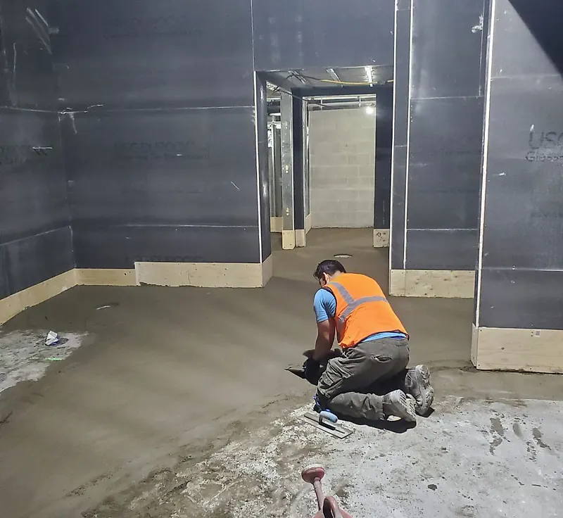 Worker in an orange hi-vis vest kneeling and floating a fresh mortar bed in a room lined with cement board