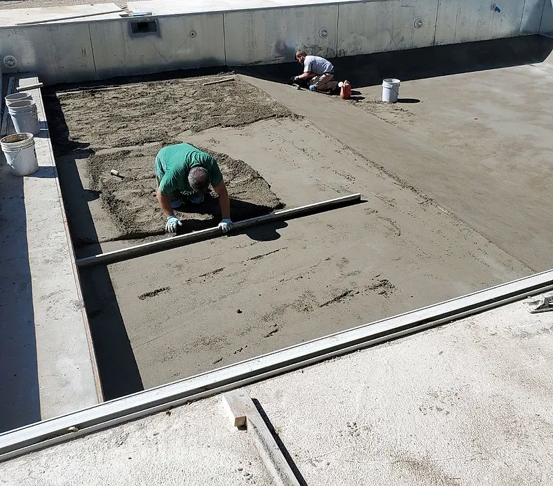 Worker kneeling with a straightedge on a fresh mortar bed on the floor of an outdoor pool, a second worker behind