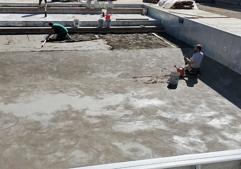 Outdoor pool floor freshly floated, one worker at the steps with a straightedge and another kneeling by a bucket